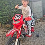 Logan participe au concours pour gagner de l'argent avec cette photo : child, toy_motorcycle, red, thumbs_up, outdoor, gravel, greenery, storage_box, curly_hair, sneakers, long_sleeve_shirt, smiling, playtime, daylight, training_wheels, number_119, pants, fun, happy, posing