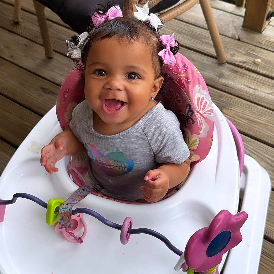 Naeliya joined the competition — help win amazing prizes! baby, child, clothing, cute, expression, face, hair_bows, happy, infant, joy, outdoor, person, pink, playful, seat, smiling, walker, white, wicker_chair, wooden_deck