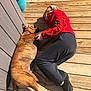 person, dog, wooden_deck, sunlight, red_jacket, black_pants, black_socks, brindle_dog, plant_pot, outdoor, relaxation, pet, companion, resting, casual_clothing, summer, shadow, natural_light, flooring, friendship