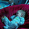 child, toddler, sleeping, baby, pajamas, couch, indoor, portrait, face, blond_hair, closed_eyes, relaxed, lounge, home, furniture, red_couch, blue_lighting, baby_supplies, star_decoration, nap