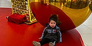 Livio participe au concours pour gagner de l'argent avec cette photo : black_pants, boy, child, christmas_decorations, curious_expression, festive, floor, golden_ball, holiday_lights, indoor, large_ornament, people, public_space, red_mat, reflection, reflection_ball, shoes, shopping_mall, sitting, winter_jacket