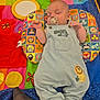 baby, sleeping, pacifier, overalls, animal_patch, colorful_quilt, pillow, patterned_pillow, child, infant, cute, resting, soft_toys, playmat, indoor, footwear, sock, hand, face, peaceful