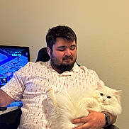 Maverick joined the competition — help win amazing prizes! man, cat, white_cat, fluffy_cat, indoor, chair, tshirt, facial_hair, earbuds, watch, television, game, relaxed, pet, human, seated, comfort, technology, casual, smile