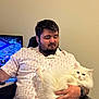 man, cat, white_cat, fluffy_cat, indoor, chair, tshirt, facial_hair, earbuds, watch, television, game, relaxed, pet, human, seated, comfort, technology, casual, smile