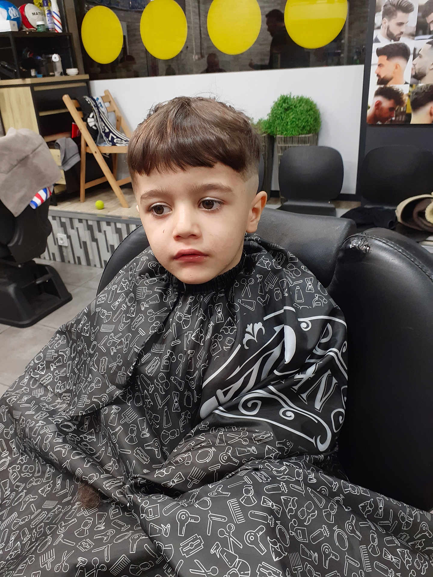 Richard participe au concours pour gagner de l'argent avec cette photo : child, boy, haircut, barber_chair, barber, salon, haircut_cape, bangs, short_hair, portrait, indoor, plant, poster, mirror, seat, expression_unimpressed, waiting, furniture, styling_tool_pattern, floor_tiles