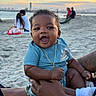 baby, child, smile, beach, sand, sunset, water, person, tattoo, necklace, curly_hair, blue_shirt, socks, holding, happy, outdoor, people, background, relaxation, nature