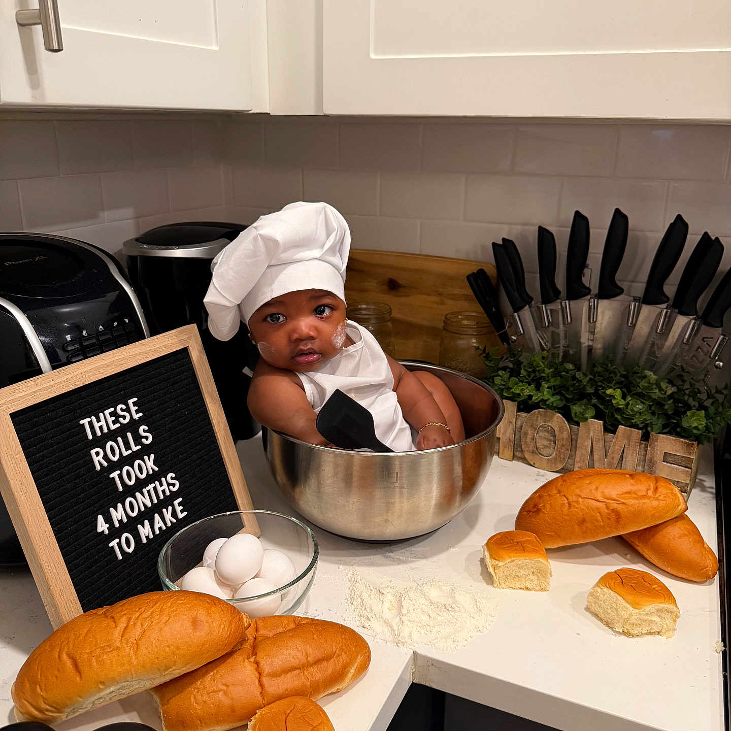 Ryaire is registered to the contest to win money with this photo: baby, chef_hat, mixing_bowl, bread, eggs, flour, kitchen_counter, kitchen, sign_board, knife_set, plant, home_decor, cute, infant, playful, cooking, food, portrait, indoors, smiling