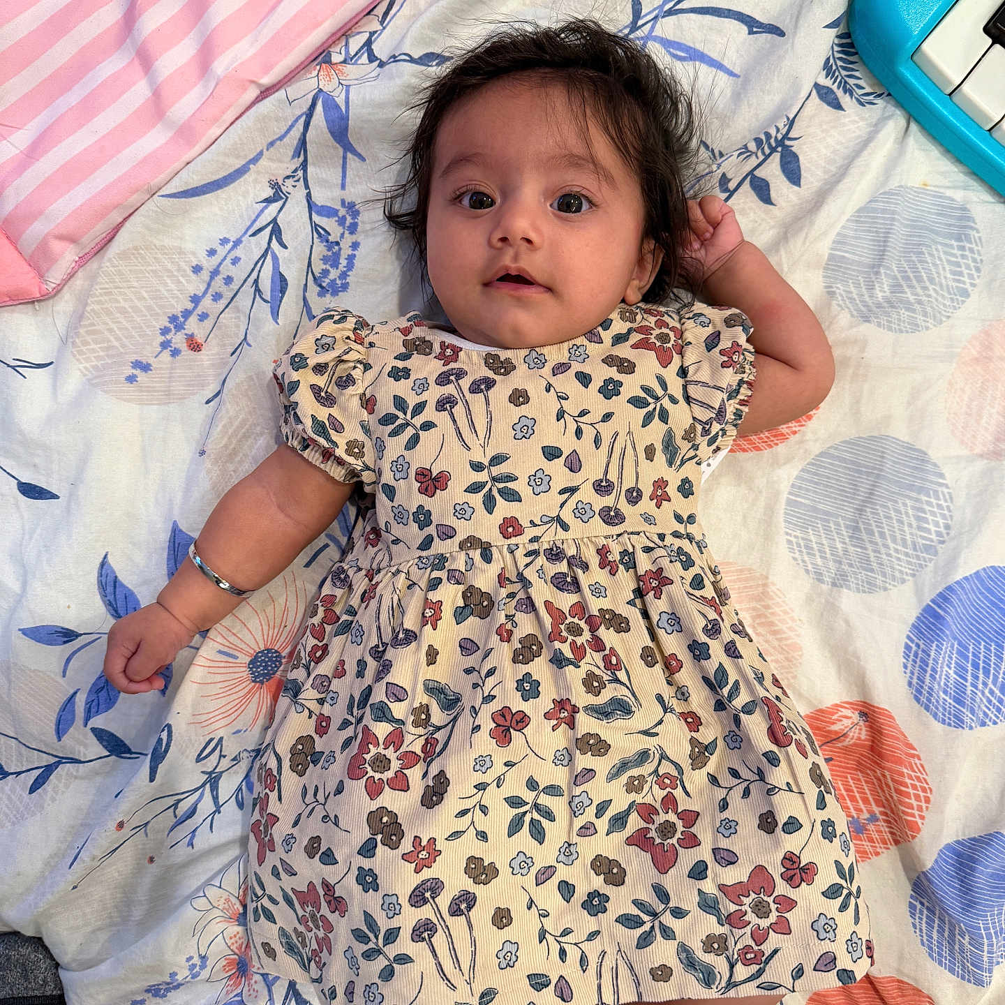 Adab joined the competition — help win amazing prizes! baby, bedspread, child, colorful, cozy, cute, floral_dress, foot, hand_holding, home, indoor, infant, lying_down, pattern, person, playful, portrait, smiling, soft_texture, toy_piano