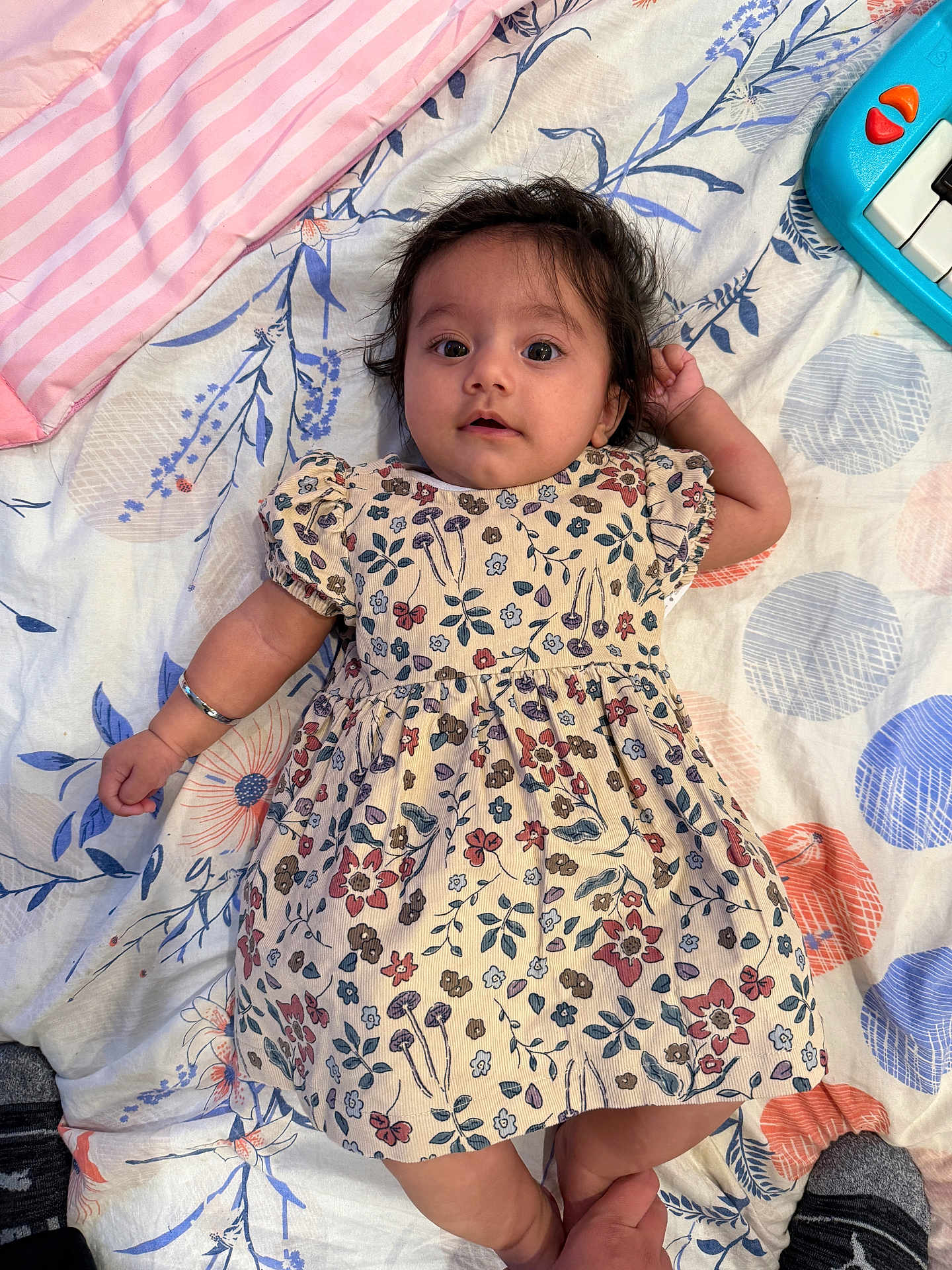 Adab joined the competition — help win amazing prizes! baby, child, infant, floral_dress, bedspread, toy_piano, lying_down, hand_holding, foot, pattern, colorful, indoor, cute, portrait, person, smiling, cozy, soft_texture, home, playful