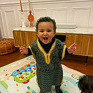 Amina participe au concours pour gagner de l'argent avec cette photo : baby, black_socks, child, colorful_toys, curly_hair, decorative_candles, floor, furniture, happy, indoor, living_room, orange_pajamas, person, play_mat, smiling, standing, toddler, traditional_clothing, wooden_cabinet