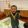 baby, black_socks, child, colorful_toys, curly_hair, decorative_candles, floor, furniture, happy, indoor, living_room, orange_pajamas, person, play_mat, smiling, standing, toddler, traditional_clothing, wooden_cabinet