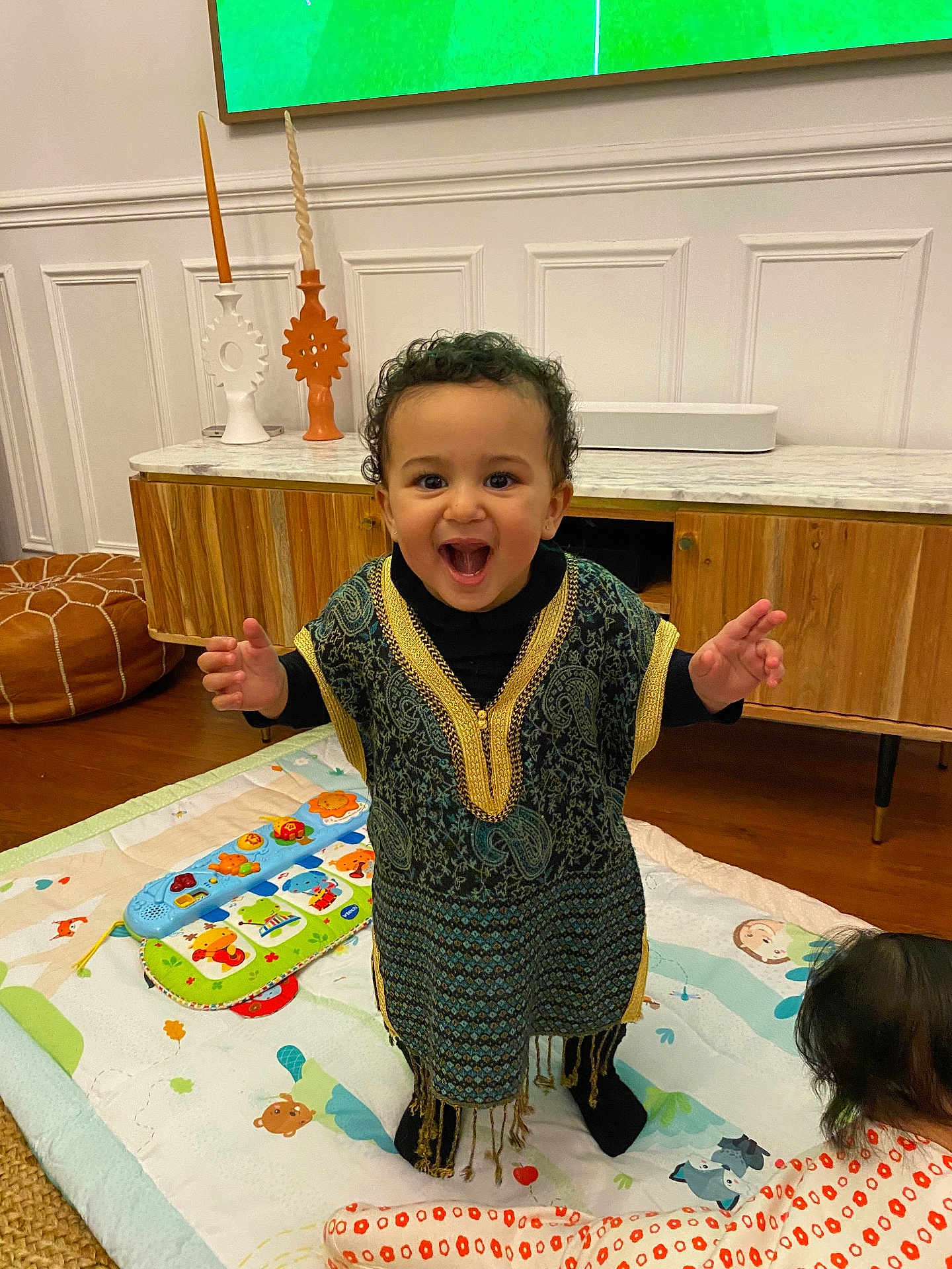 Amina participe au concours pour gagner de l'argent avec cette photo : baby, black_socks, child, colorful_toys, curly_hair, decorative_candles, floor, furniture, happy, indoor, living_room, orange_pajamas, person, play_mat, smiling, standing, toddler, traditional_clothing, wooden_cabinet