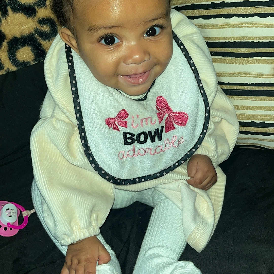 Kmiyah is registered to the contest to win money with this photo: baby, child, infant, smiling, pigtails, bib, clothing, sitting, cushion, striped_pillow, leopard_print, hand, footed_pajamas, black_background, cute, indoors, portrait, happy, person, young_child