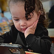 Azalea is registered to the contest to win money with this photo: toddler, child, curly_hair, smartphone, black_sweater, hand_on_face, indoor, table, blurred_background, focused, portrait, young_child, casual_clothing, seated, cafe, cute, technology, concentration, person, face