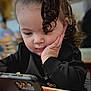 toddler, child, curly_hair, smartphone, black_sweater, hand_on_face, indoor, table, blurred_background, focused, portrait, young_child, casual_clothing, seated, cafe, cute, technology, concentration, person, face