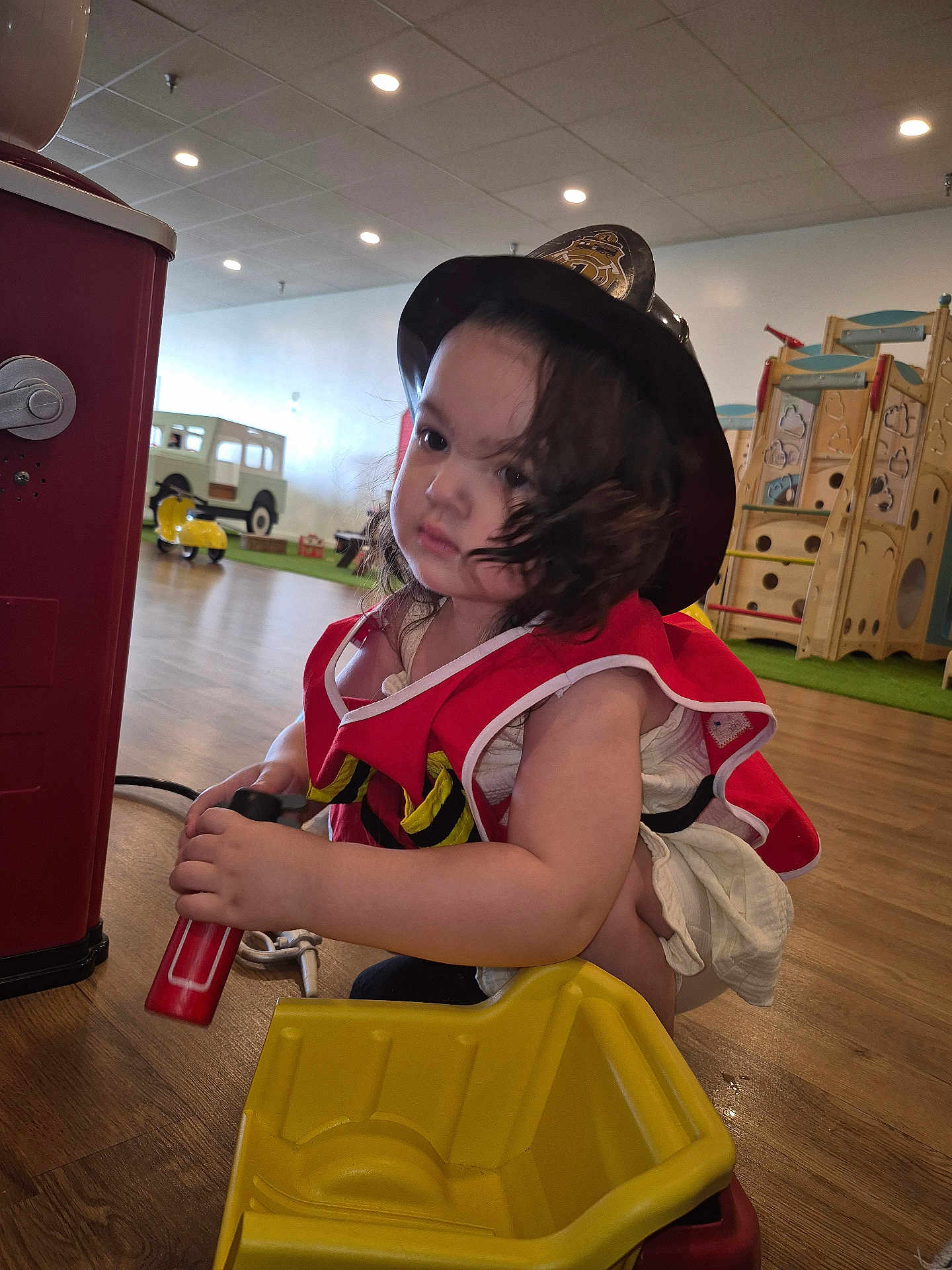 Azalea is registered to the contest to win money with this photo: child, toddler, indoor, playground, toy, firefighter_costume, helmet, red_vest, yellow_toy, wooden_floor, play_equipment, curly_hair, crouching, young_child, hands, face, expression, play_area, bright_lighting, casual_clothing
