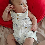 Caelum is registered to the contest to win money with this photo: baby, infant, child, romper, clothing, red_cushion, bed, fabric, skin, hand, face, cute, portrait, indoor, relaxing, gazing, blue_eyes, sitting, newborn, soft