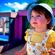Manar is registered to the contest to win money with this photo: toddler, child, sunhat, blue_sky, outdoor, daylight, smiling, yellow_clothing, floral_pattern, playground, wooden_deck, happy, cute, portrait, summer, person, baby, curly_hair, sunlight, nature
