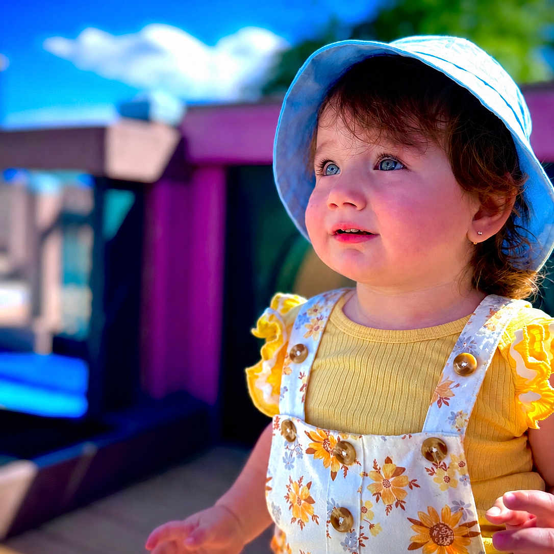 Manar is registered to the contest to win money with this photo: toddler, child, sunhat, blue_sky, outdoor, daylight, smiling, yellow_clothing, floral_pattern, playground, wooden_deck, happy, cute, portrait, summer, person, baby, curly_hair, sunlight, nature