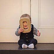 Asher is registered to the contest to win money with this photo: baby, cardboard, checkered_shirt, child, cute, doorstep, face, front_door, indoor, letter_o, neutral_background, playful, portrait, prop, sitting, smile, socks, thumbs_up, toddler, vest