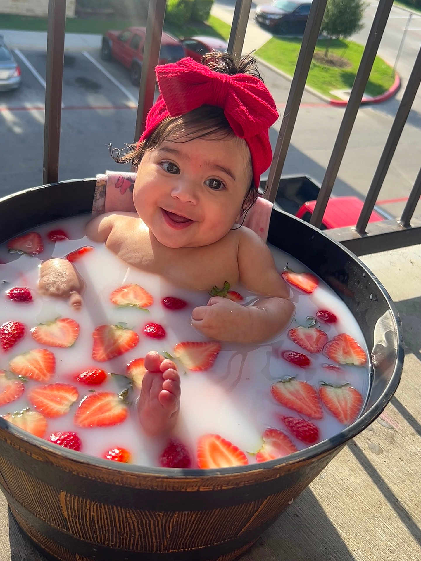 Audrey is registered to the contest to win money with this photo: baby, child, milk_bath, strawberries, red_bow, headband, wooden_tub, outdoor, sunlight, smiling, happy, foot, water, fruit, balcony, railing, concrete_floor, parked_cars, greenery, person