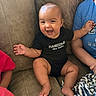 baby, child, couch, smiling, happy, black_onesie, clothing, person, family, indoor, furniture, arm, hand, foot, blanket, blue_shirt, skin, sitting, laughing, baby_face