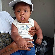 Josiah is registered to the contest to win money with this photo: baby, child, cap, drool, tattoo, hand, person, sleeveless_shirt, blue_shorts, indoor, wall, stroller, curious, holding, clothing, face, portrait, infant, skin, expression