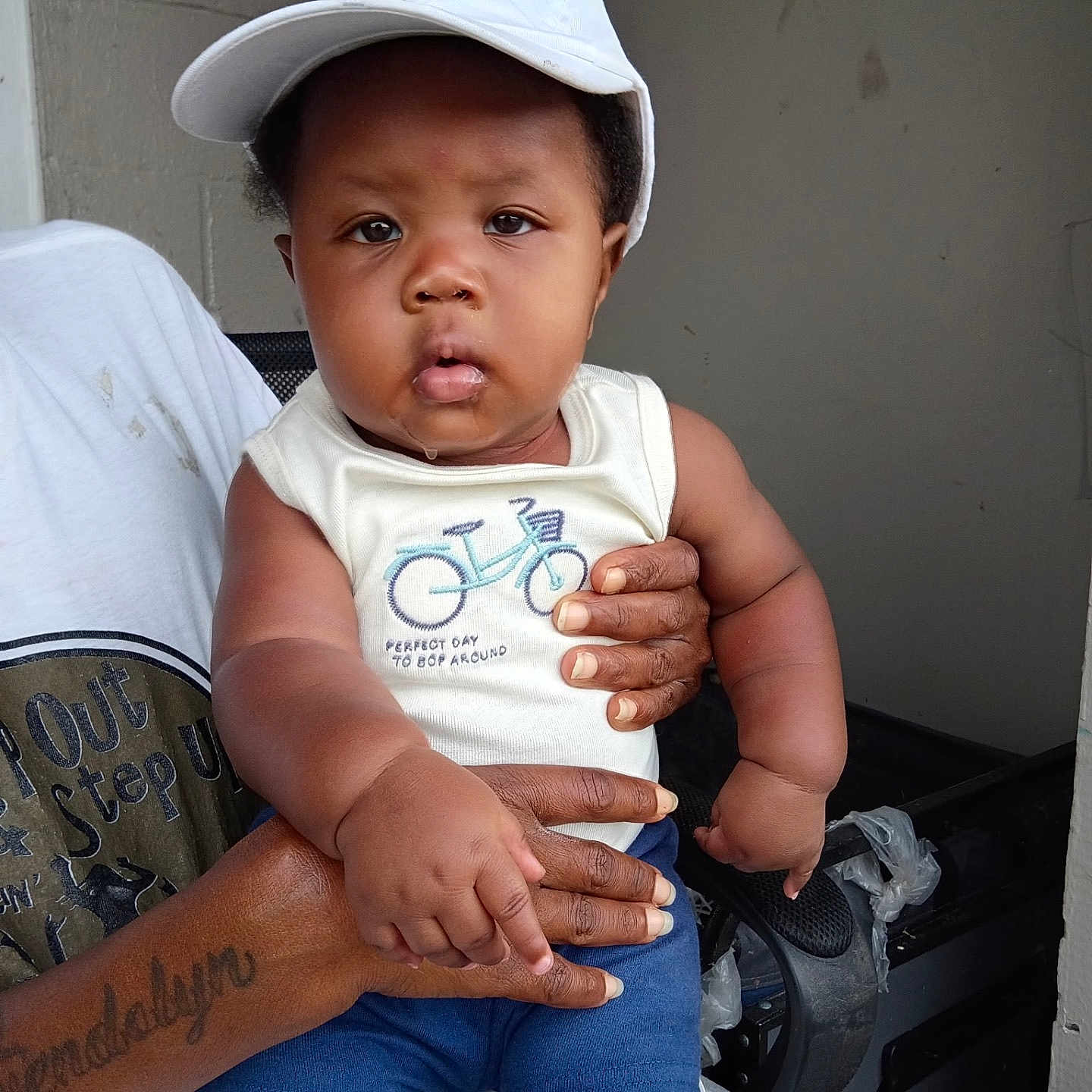 Josiah is registered to the contest to win money with this photo: baby, child, cap, drool, tattoo, hand, person, sleeveless_shirt, blue_shorts, indoor, wall, stroller, curious, holding, clothing, face, portrait, infant, skin, expression