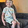 child, smiling, curly_hair, tshirt, shorts, boots, sitting, couch, indoor, happy, casual_clothing, footwear, furniture, young_child, portrait, home, relaxed, cute, person, cozy