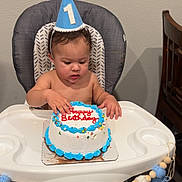 Noah is registered to the contest to win money with this photo: baby, birthday_cake, high_chair, party_hat, blue_icing, sprinkles, tassels, banner, number_one, celebration, child, indoor, table, wooden_chair, face, hand, skin, wall, floor, clothing