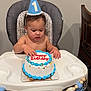 baby, birthday_cake, high_chair, party_hat, blue_icing, sprinkles, tassels, banner, number_one, celebration, child, indoor, table, wooden_chair, face, hand, skin, wall, floor, clothing