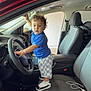 child, toddler, car, steering_wheel, driver_seat, blue_shirt, checkered_shorts, sneakers, interior, vehicle, curly_hair, standing, door_open, dashboard, floor_mat, car_seat, seatbelt, hand, looking, garage