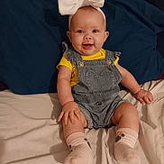 Hope is registered to the contest to win money with this photo: baby, child, smiling, headband, bow, denim_overalls, yellow_shirt, sitting, bed, pillow, happy, cute, infant, sock, clothing, person, indoor, portrait, skin, young