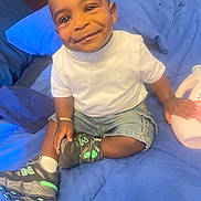Josiah joined the competition — help win amazing prizes! toddler, child, smile, indoor, bed, blue_bedspread, white_tshirt, denim_shorts, sneakers, happy, cute, person, young_child, casual_clothing, seated, bracelet, toy, comfort, portrait, candid