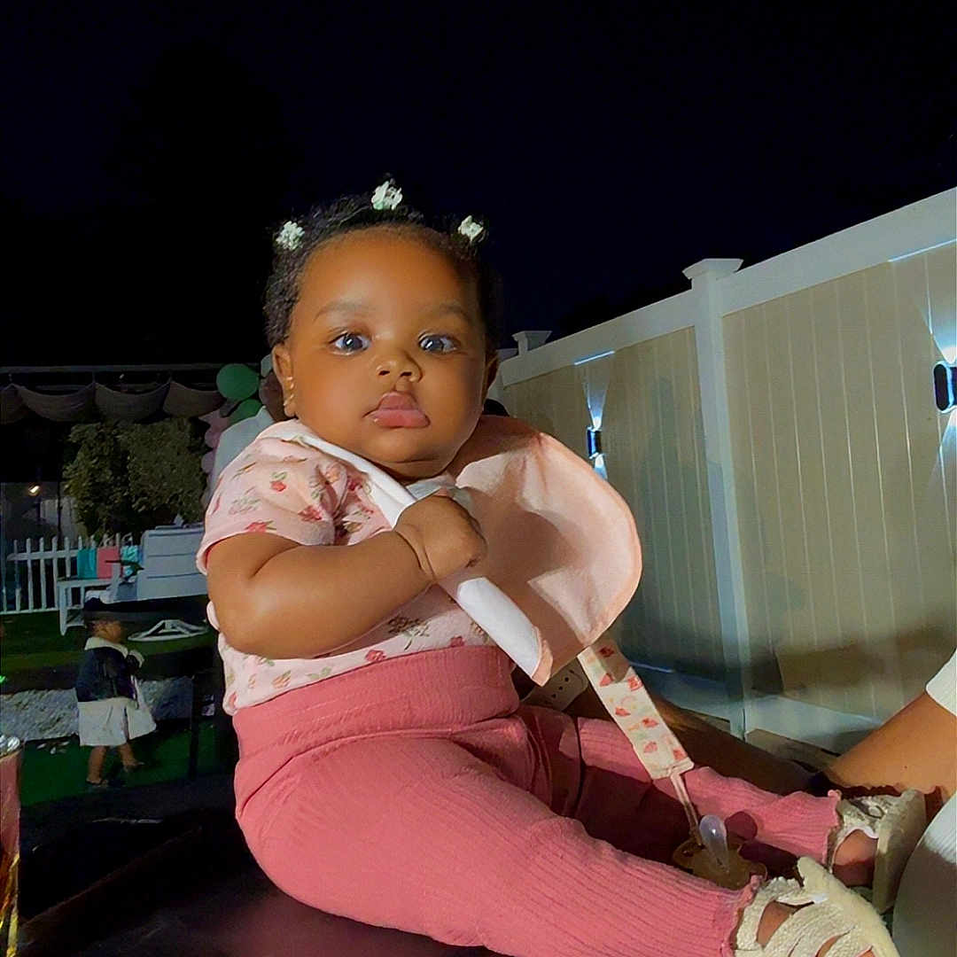 Zaira Kerène Victor is registered to the contest to win money with this photo: baby, child, infant, portrait, sitting, outdoor, night, backyard, playground, pink_clothing, leggings, shoes, hair_clips, flower_accessories, serious_expression, flash_photography, fence, party, play_equipment, family_photo