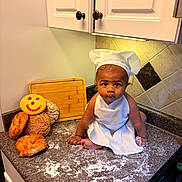 Iván is registered to the contest to win money with this photo: baby, chef_hat, apron, kitchen_counter, bread, cookie, smiley_face, flour, cutting_board, tile_wall, cabinet, baby_boy, cooking, baking, food, portrait, cute, indoor, child, kneeling