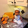 baby, chef_hat, apron, kitchen_counter, bread, cookie, smiley_face, flour, cutting_board, tile_wall, cabinet, baby_boy, cooking, baking, food, portrait, cute, indoor, child, kneeling