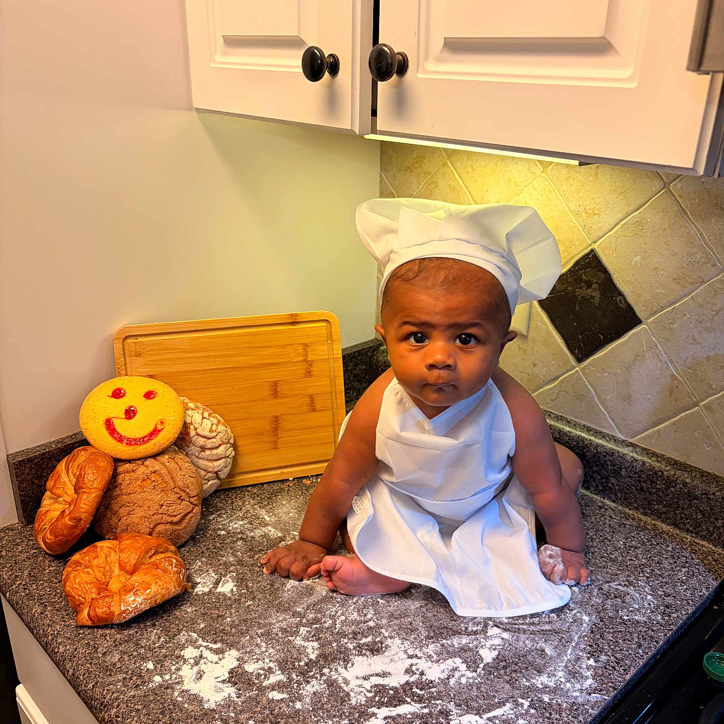 Iván is registered to the contest to win money with this photo: baby, chef_hat, apron, kitchen_counter, bread, cookie, smiley_face, flour, cutting_board, tile_wall, cabinet, baby_boy, cooking, baking, food, portrait, cute, indoor, child, kneeling