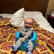 Elijah joined the competition — help win amazing prizes! baby, child, smiling, blue_baby_seat, bed, blanket, pillow, gray_outfit, purple_socks, wooden_headboard, indoor, remote_control, bedroom, furniture, person, home, happy, sitting, young_child, portrait