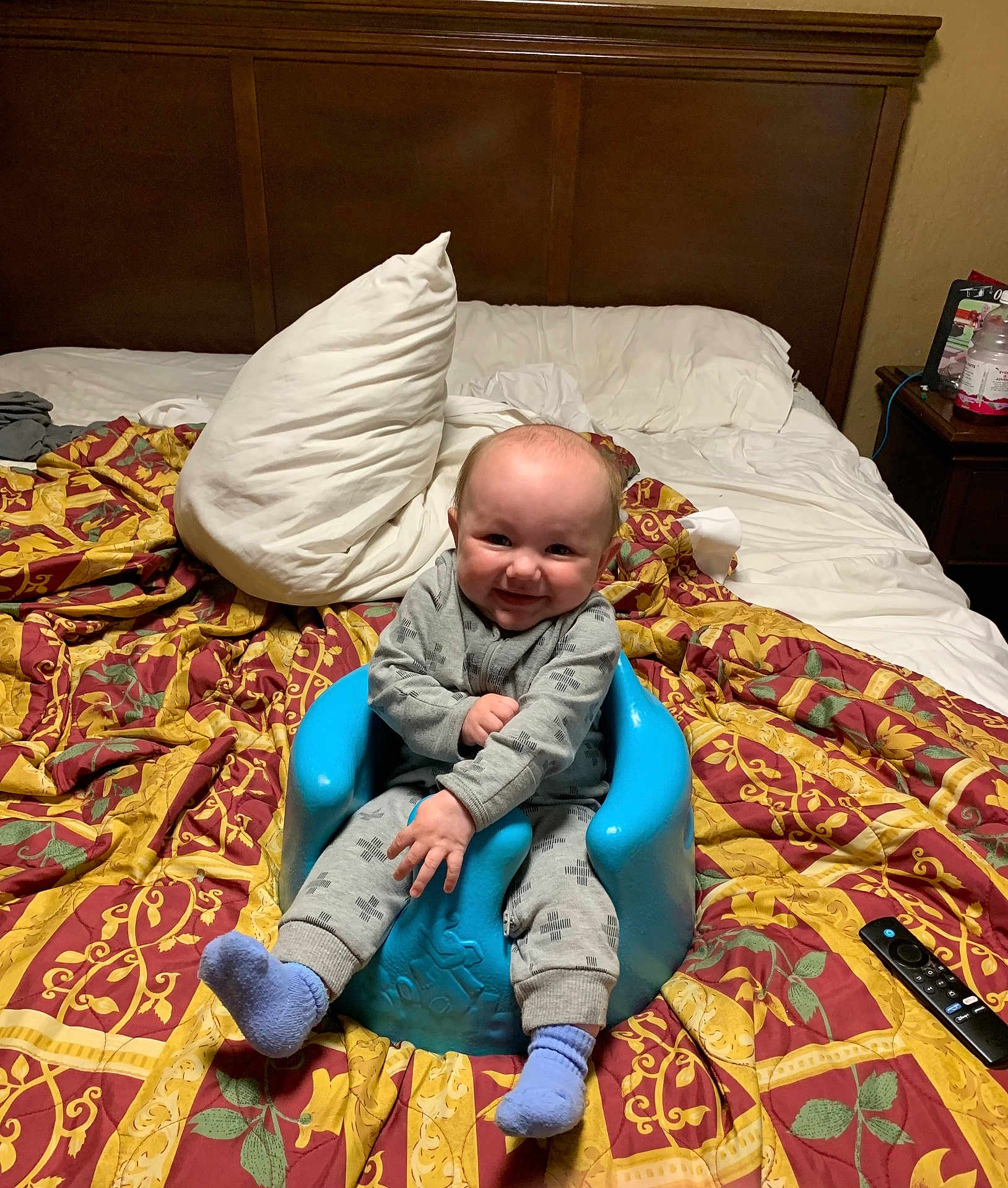 Elijah joined the competition — help win amazing prizes! baby, child, smiling, blue_baby_seat, bed, blanket, pillow, gray_outfit, purple_socks, wooden_headboard, indoor, remote_control, bedroom, furniture, person, home, happy, sitting, young_child, portrait