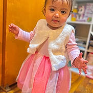 Eliana is registered to the contest to win money with this photo: toddler, child, girl, dress, pink, vest, indoor, wooden_floor, hair_clips, barefoot, curious_expression, standing, sleeves, home, shelf, toy, young_child, cute, portrait, person