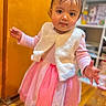 barefoot, child, curious_expression, cute, dress, girl, hair_clips, home, indoor, person, pink, portrait, shelf, sleeves, standing, toddler, toy, vest, wooden_floor, young_child