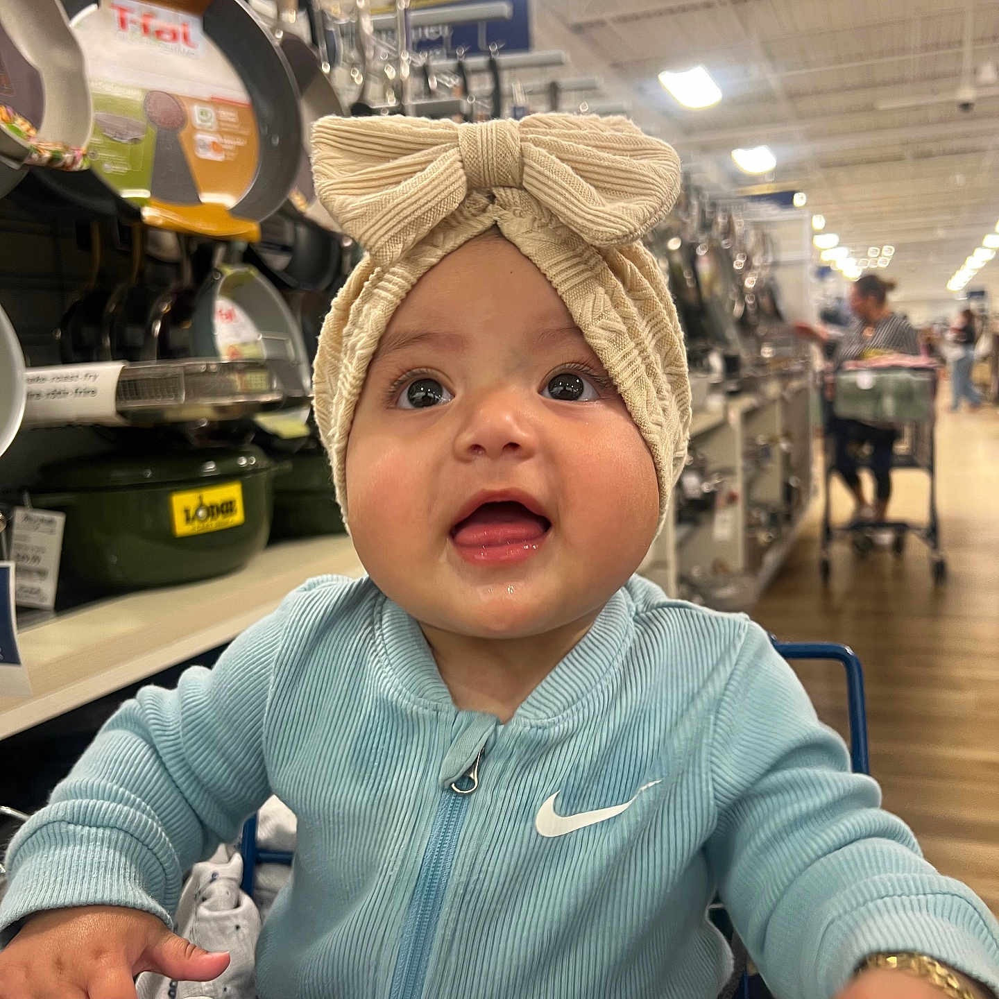 Eliana joined the competition — help win amazing prizes! aisle, baby, bow, child, clothing, curious, face, happy, headband, indoor, infant, jacket, kitchenware, pans, person, pots, retail, shopping_cart, smile, store
