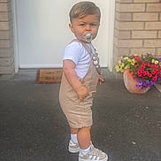 Isaac participe au concours pour gagner de l'argent avec cette photo : toddler, child, pacifier, overalls, white_shirt, sneakers, standing, driveway, door, flower_pot, flowers, brick_wall, outdoor, side_view, young_child, casual_clothing, short_hair, white_socks, serious_expression, daylight