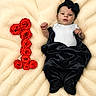 adorable, baby, baby_clothing, black_bow, black_pants, celebration, cute, flower_arrangement, fluffy_blanket, headband, infant, lying_down, milestone, newborn, number_one, portrait, red_roses, resting, soft_texture, white_onesie