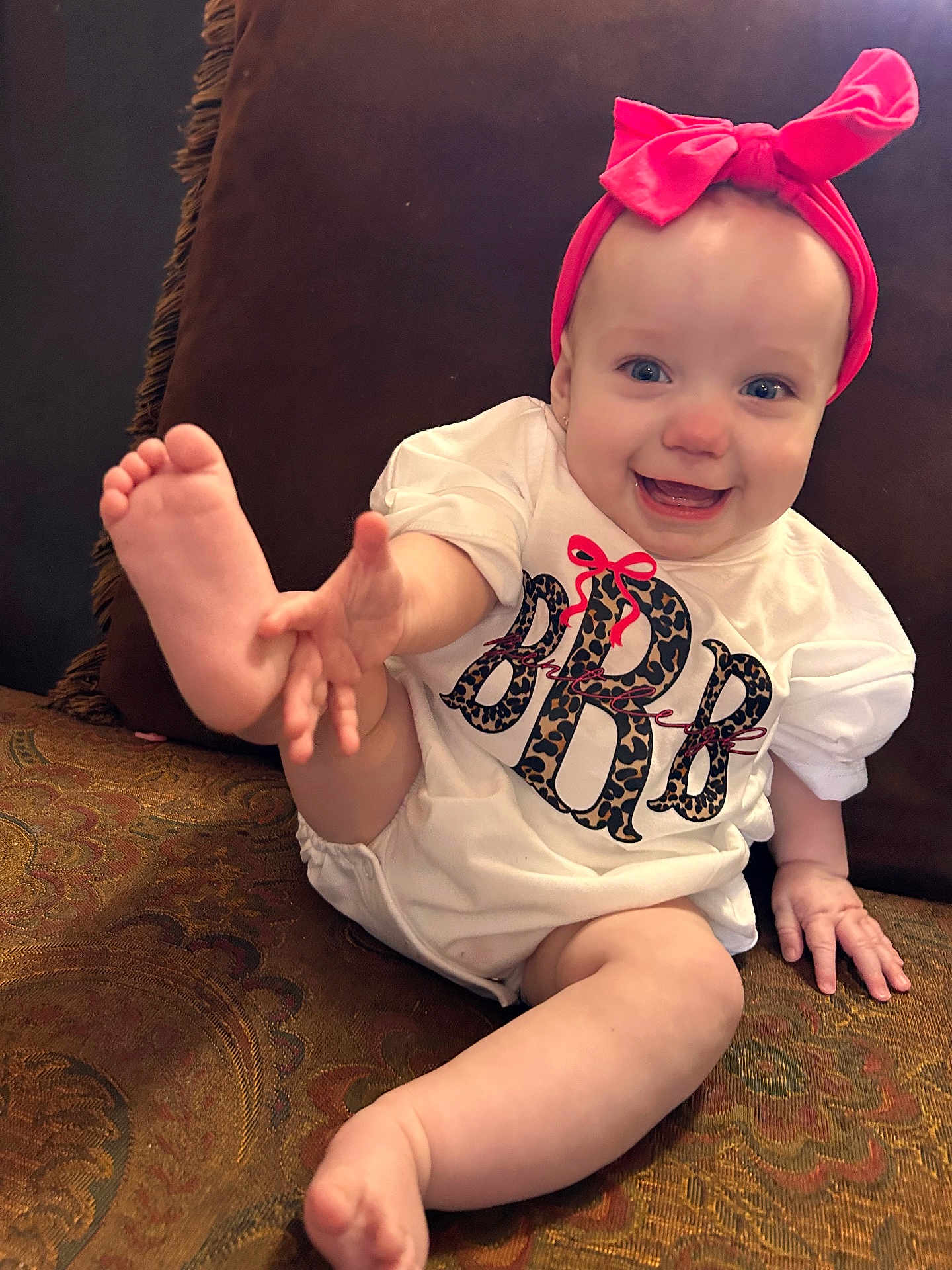 Bentleigh is registered to the contest to win money with this photo: baby, infant, child, smiling, headband, pink_bow, onesie, foot, toes, hand, couch, cushion, patterned_fabric, living_room, happy, portrait, sitting, blue_eyes, earring, cheerful
