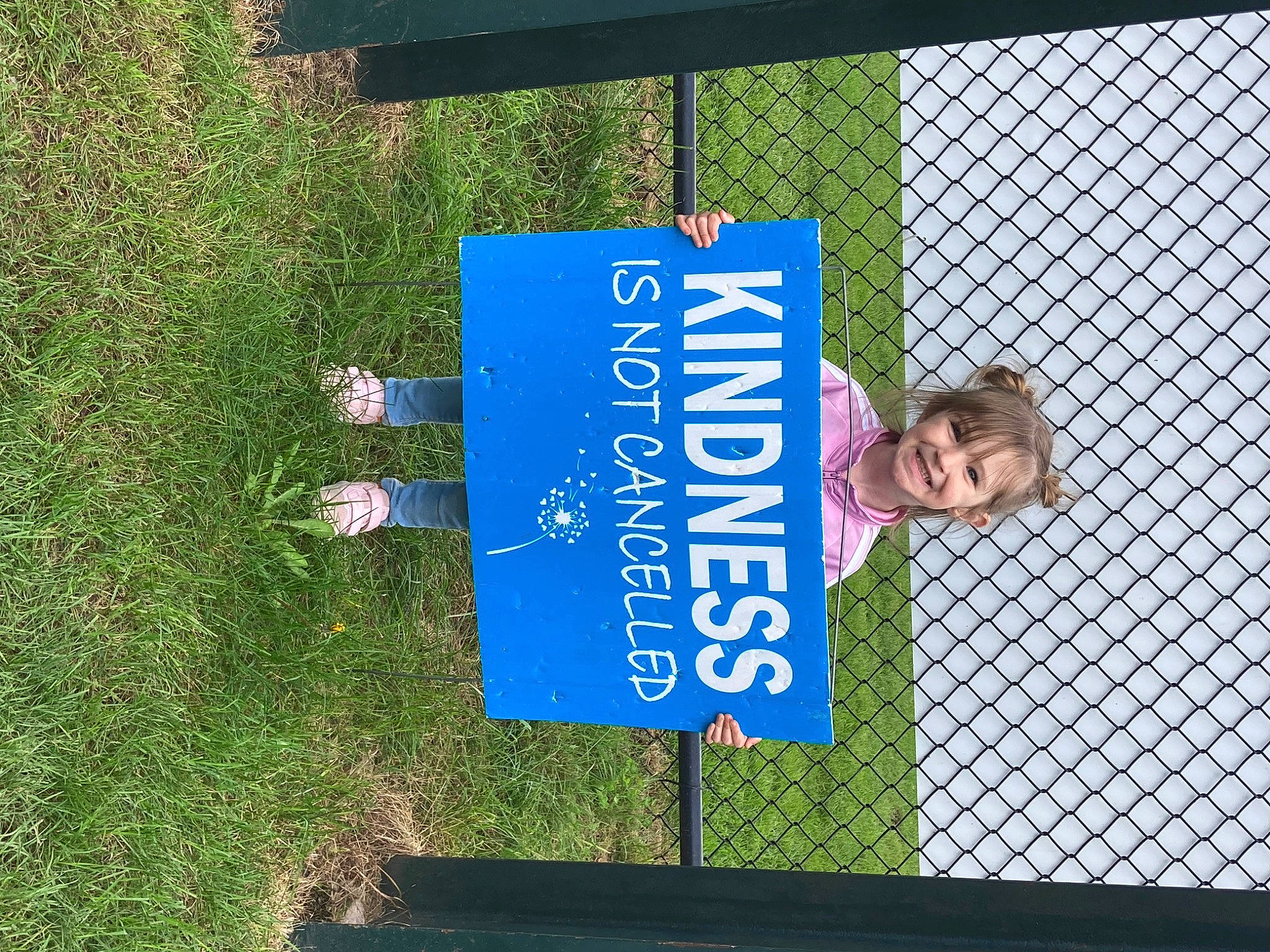 Emma is registered to the contest to win money with this photo: advertising, chain_link_fencing, electric_blue, fence, font, grass, joy, leisure, logo, mesh, metal, net, person, plant, public_utility, sign, signage, sunglasses, t_shirt, tree