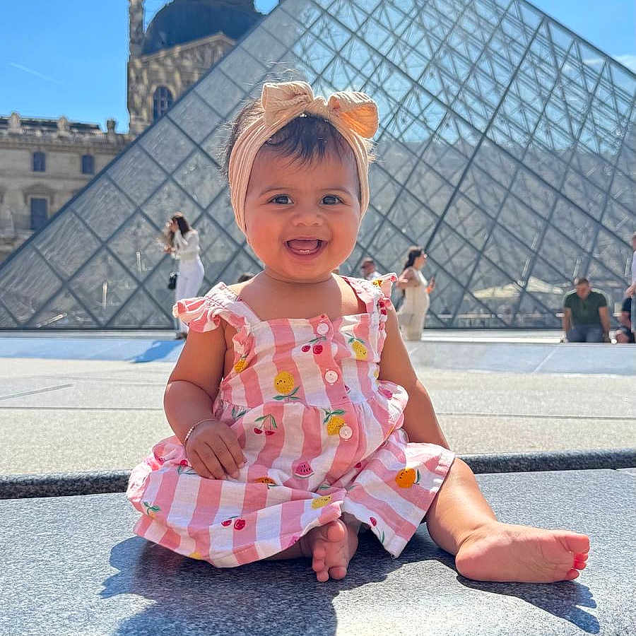 Deniya a rejoint le concours — aidez-le/la à gagner de superbes lots ! architecture, baby, building, child, daylight, dress, footwearless, glass_pyramid, happy, headband, louvre, outdoor, people, portrait, sitting, sky, smiling, stone, sunny, tourist