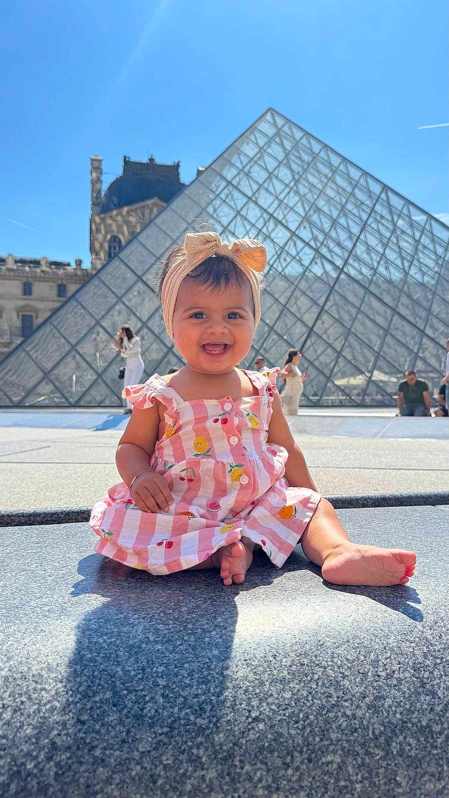 Deniya a rejoint le concours — aidez-le/la à gagner de superbes lots ! architecture, baby, building, child, daylight, dress, footwearless, glass_pyramid, happy, headband, louvre, outdoor, people, portrait, sitting, sky, smiling, stone, sunny, tourist