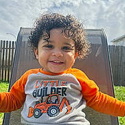 Devante' joined the competition — help win amazing prizes! toddler, child, smiling, curly_hair, outdoor, chair, sweater, orange, white, mesh_chair, fence, grass, sunny, cloudy_sky, portrait, happy, casual_clothing, young_child, daylight, backyard